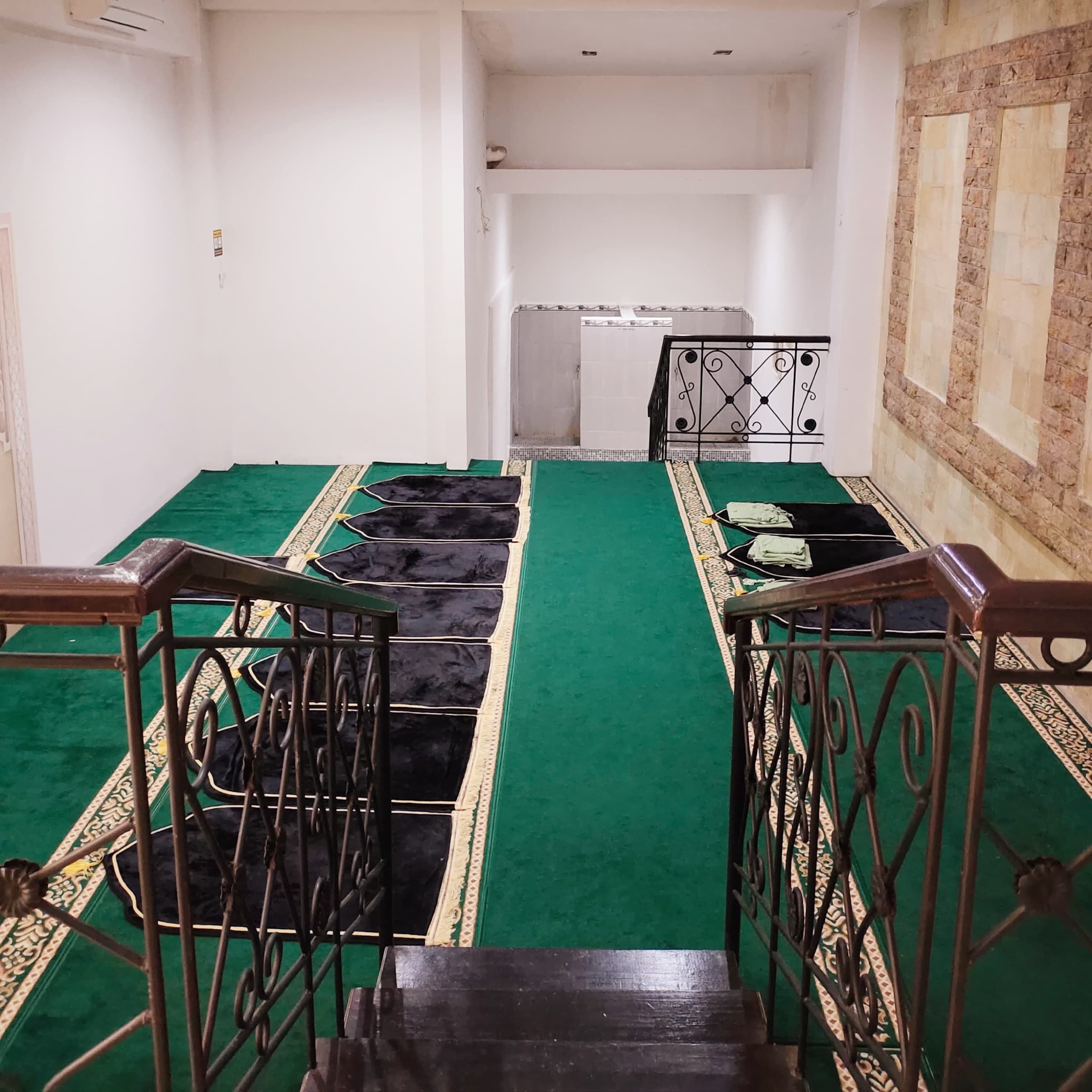 Une salle de prière avec des rangées de tapis noirs pour la prière, une moquette verte et des rampes en métal.