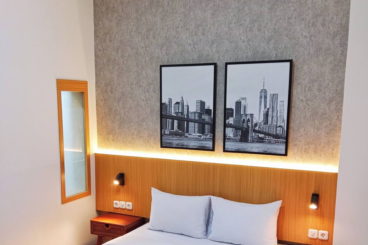 Habitación de hotel con una cama grande, dos lámparas de pared, una obra de arte con un paisaje urbano, un espejo y un equipo de aire acondicionado.