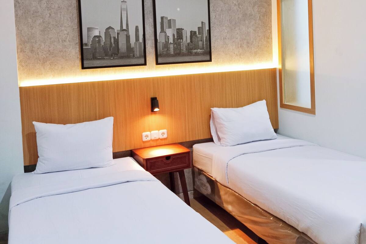 Habitación de hotel con dos camas individuales, cabecero de madera, mesita de noche con lámpara y dos cuadros con vistas urbanas en la pared.
