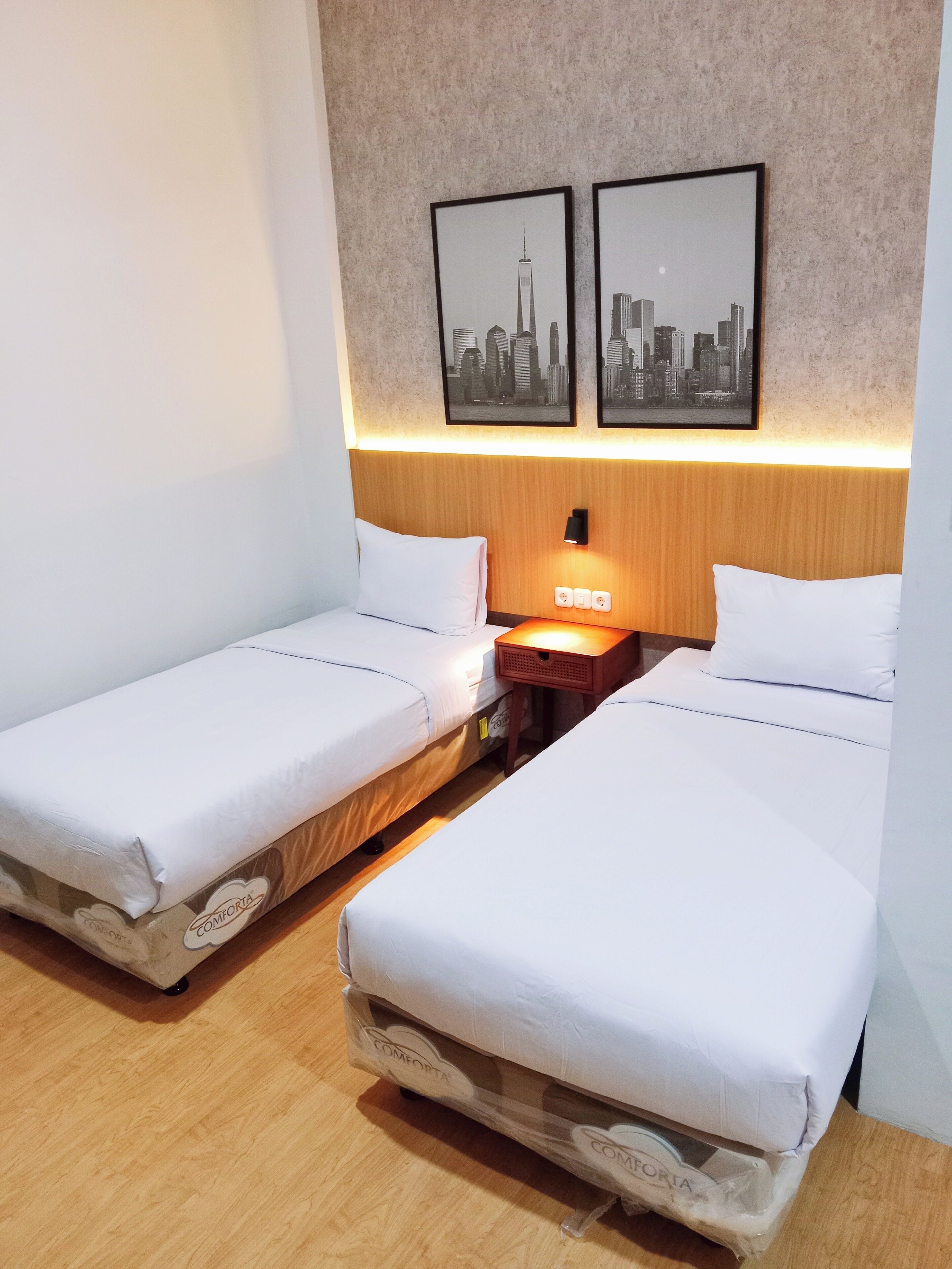 A hotel room with two single beds, a wooden headboard, a nightstand with a lamp, and two framed cityscape pictures on the wall.