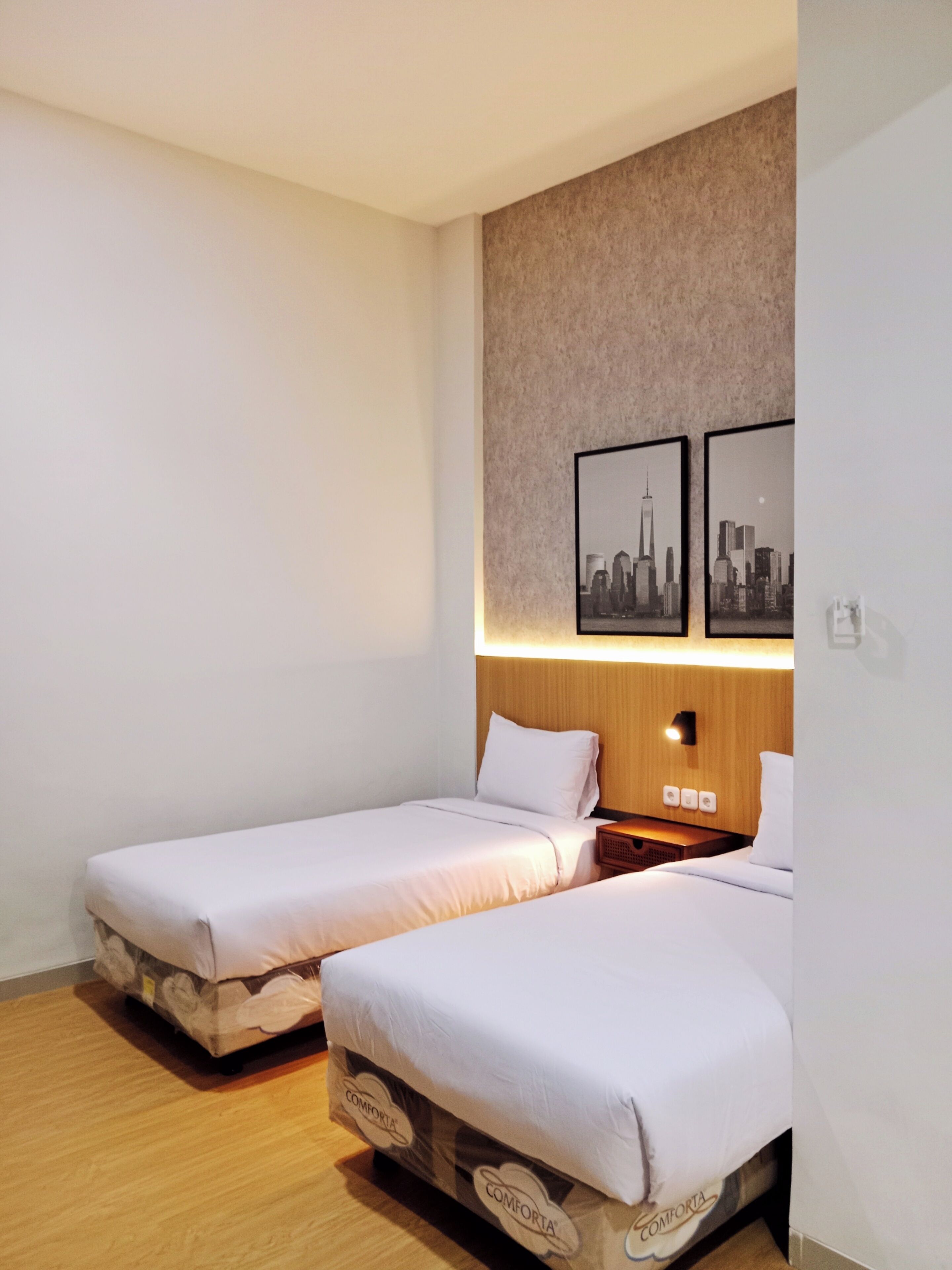 Une chambre d’hôtel avec deux lits simples, une tête de lit en bois, une table de chevet et deux tableaux encadrés représentant des vues de la ville, accrochés au mur.
