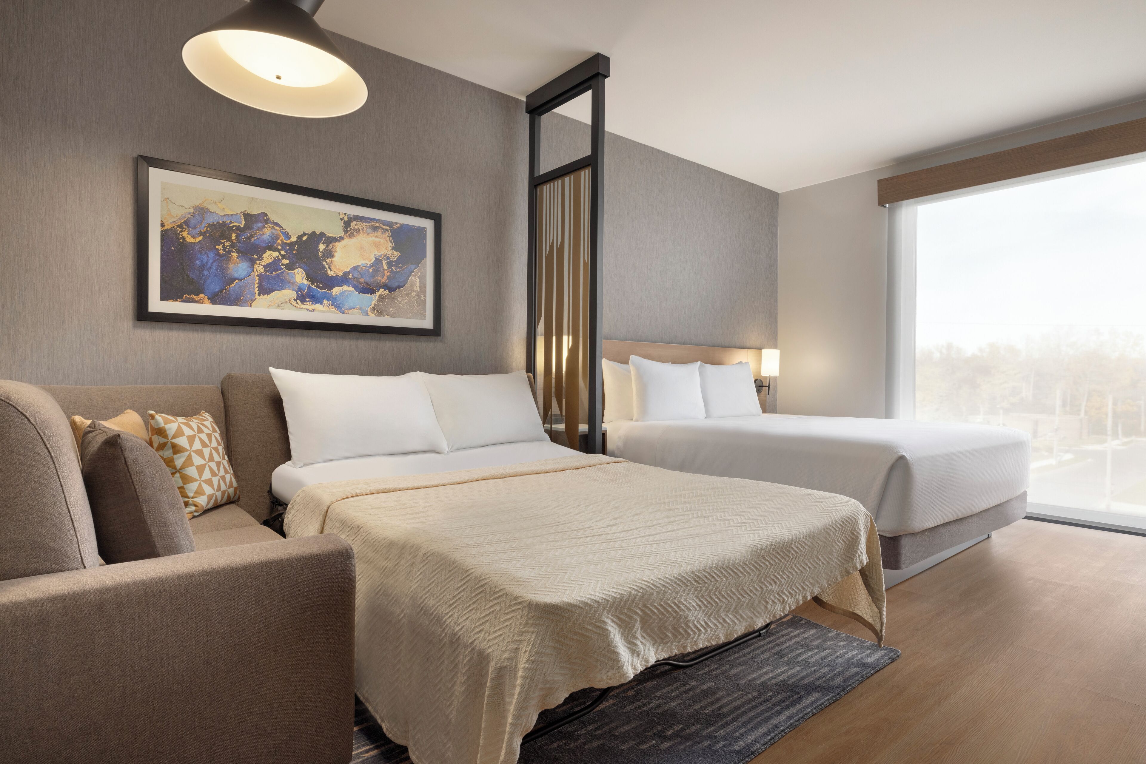 Una habitación de hotel con dos camas, un sofá, un ventanal grande y una obra de arte enmarcada en la pared.