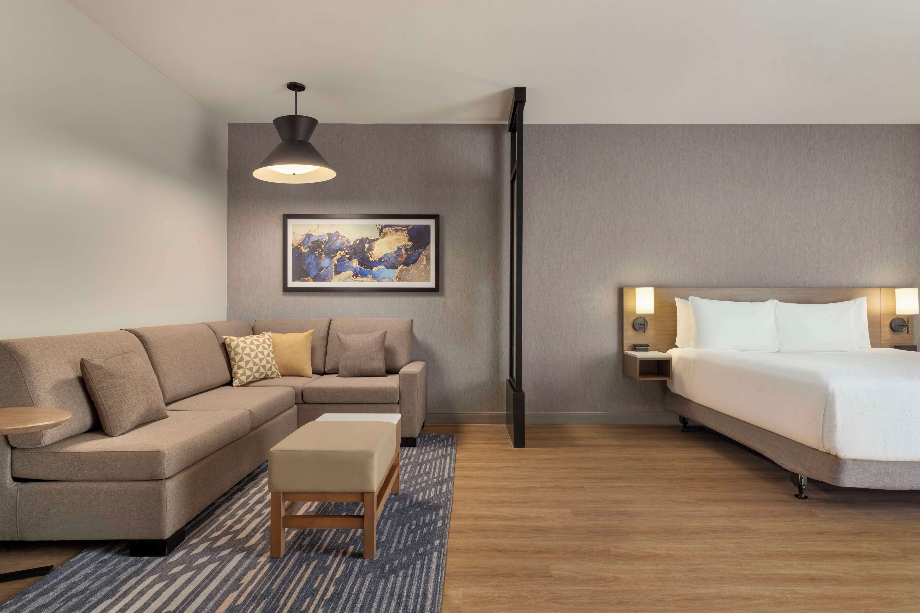 Quarto de hotel moderno com sofá, cama, mesa pequena, tapete e uma pintura na parede.