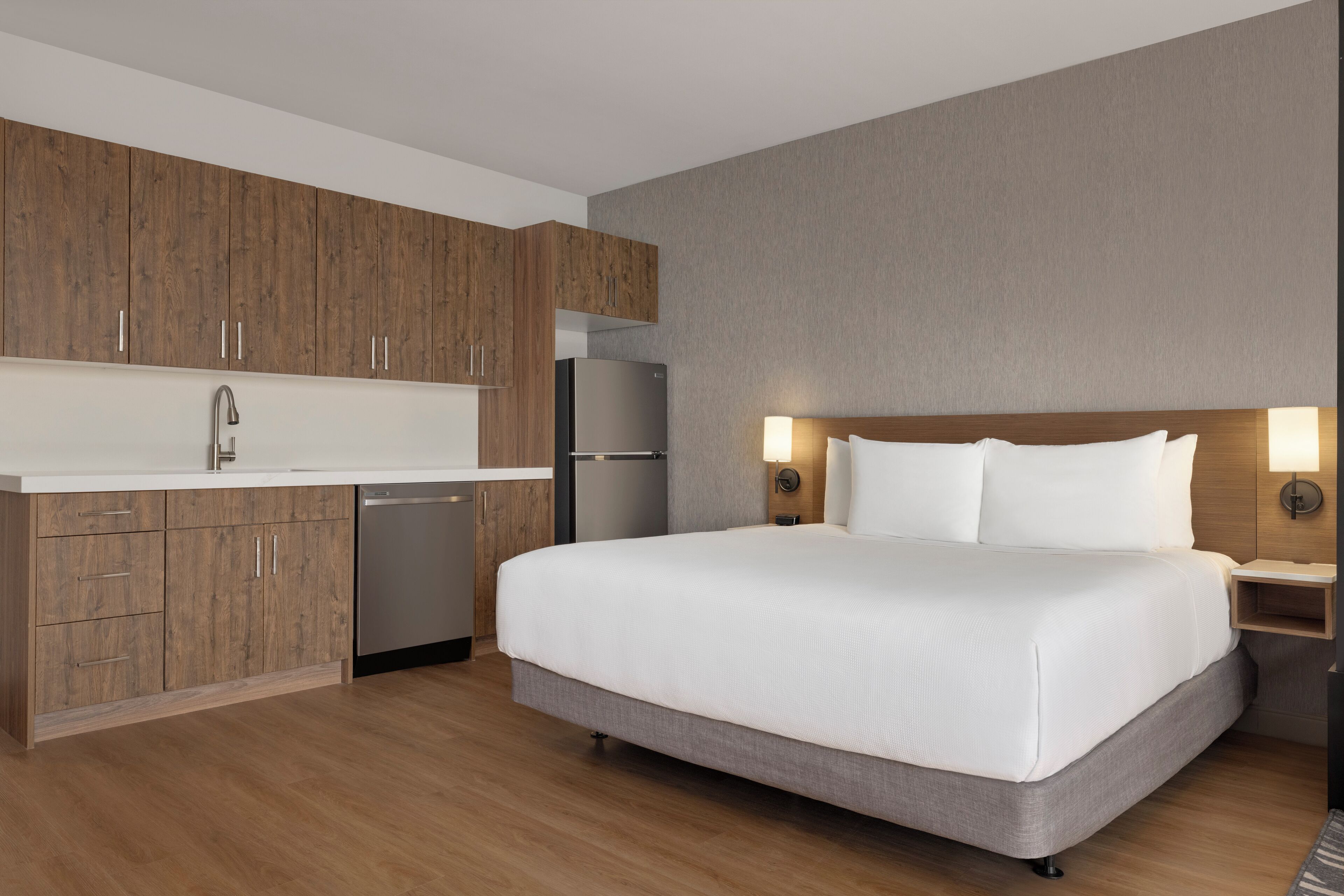 A modern hotel room with a large bed, a built-in desk, a kitchenette, and a bathroom.