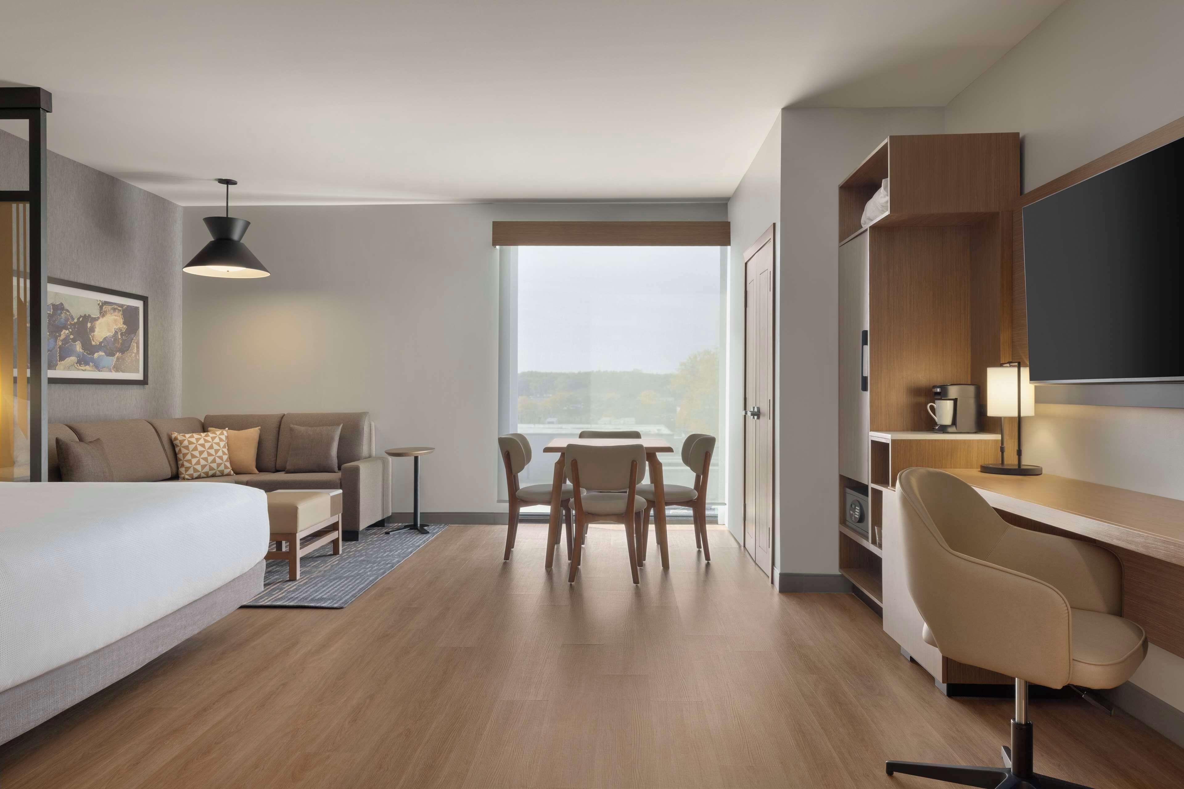 A modern hotel room with a bed, a desk, a chair, a dining table, and a large window.