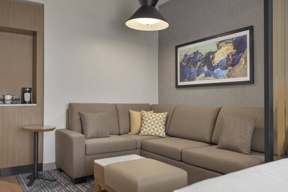 Ein modernes Hotelzimmer mit einer Eckcouch, einem kleinen runden Tisch, einer Kaffeemaschine und einem gerahmten abstrakten Gemälde an der Wand.