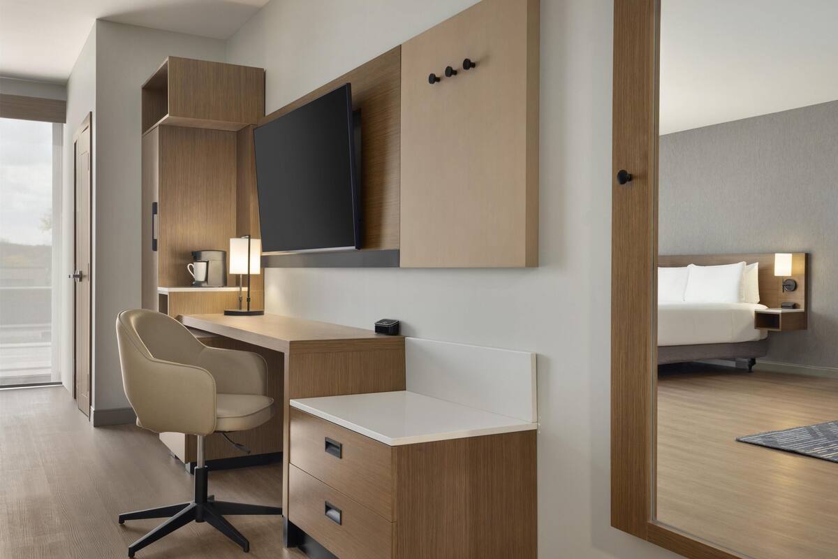 Ein modernes Hotelzimmer mit einem Holztisch, einem Sessel, einem Flachbildfernseher, einem Bett mit Kopfteil und einem Nachttisch mit Lampe.