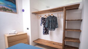 Un dormitorio con una cama, un armario de madera con ropa colgada, un banco de madera y una cómoda de madera con toallas dobladas.