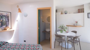 Una habitación moderna y luminosa con cama, una pequeña zona de comedor con mesa y sillas, un baño con lavamanos e inodoro, y una estantería con objetos decorativos.