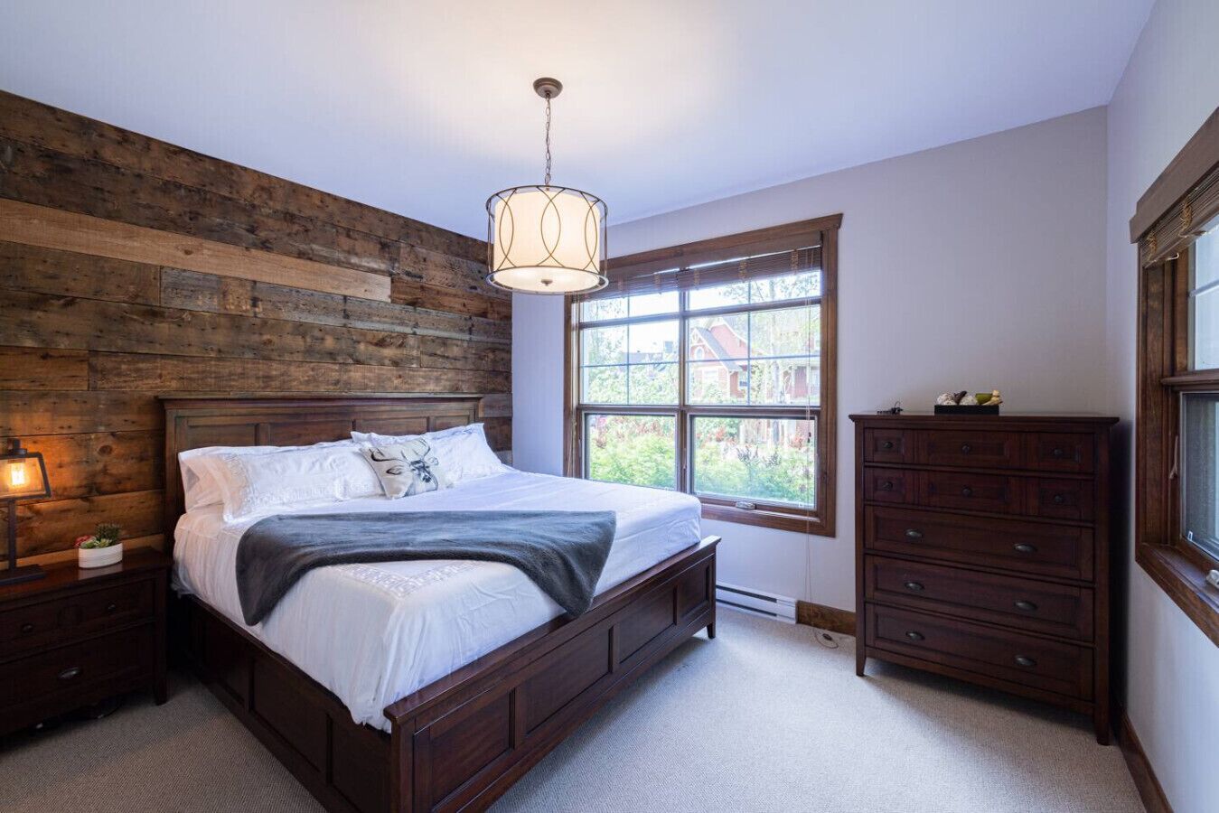 Une chambre à coucher avec une tête de lit en bois, une grande fenêtre, une commode et un luminaire suspendu.