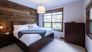 A bedroom with a wooden headboard, a large window, a dresser, and a hanging light fixture.