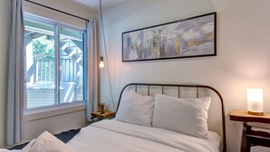 A bedroom with a bed, a window with a view of a deck, a painting on the wall, and a bedside lamp. - Cozy Lodge in the Mont Tremblant Ski Village (Mont-Tremblant)