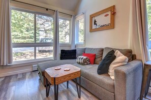Ein gemütliches Wohnzimmer mit grauem Sofa, einem kleinen Holztisch und einem großen Fenster mit Blick auf Bäume und eine Terrasse.