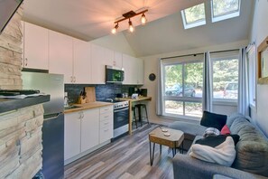 A modern kitchen with white cabinets, a built-in microwave, and a stove. There is a dining area with a gray sofa and a small table. Large windows provide natural light and a view of the outside.