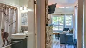 A bathroom with a sink, mirror, and shower curtain, a stone wall, a seating area with a sofa and table, and a TV mounted on the wall. - Cozy Lodge in the Mont Tremblant Ski Village (Mont-Tremblant)
