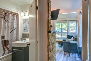 A bathroom with a sink, mirror, and shower curtain, a stone wall, a seating area with a sofa and table, and a TV mounted on the wall. - Cozy Lodge in the Mont Tremblant Ski Village (Mont-Tremblant)