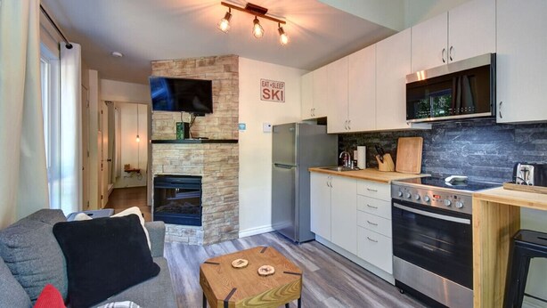 A modern kitchen with a stone fireplace, a built-in oven, and a dining area with a small table and sofa. - Cozy Lodge in the Mont Tremblant Ski Village (Mont-Tremblant)