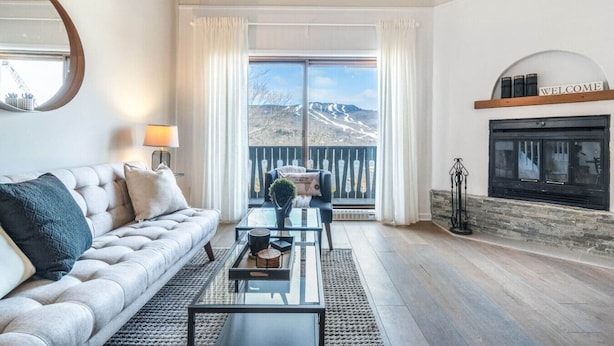 A spacious living room with a sofa, coffee table, and fireplace. A large window offers a view of a mountainous landscape. - Bright & Modern Lac Tremblant Condo + Views (Mont-Tremblant)