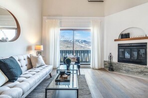 A spacious living room with a sofa, coffee table, and fireplace. A large window offers a view of a mountainous landscape.