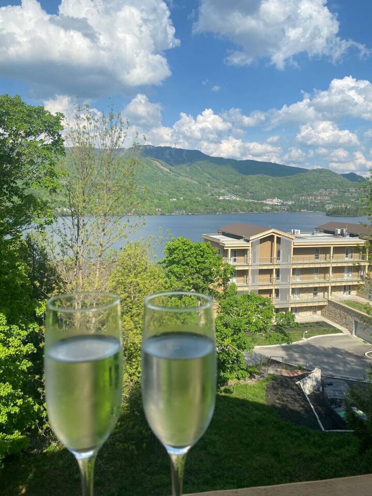 Due bicchieri d'acqua su un balcone con vista su un lago, montagne e un edificio con diversi piani e balconi.
