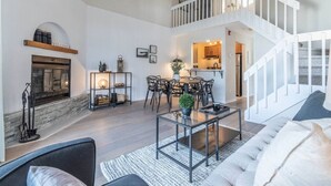 A spacious living area with a fireplace, a dining table, and a staircase leading to an upper level. - Bright & Modern Lac Tremblant Condo + Views (Mont-Tremblant)