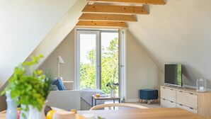 Ett modernt sovrum i en loftvåning med ett sluttande tak, ett stort fönster, en säng, en tv och en matplats med ett bord och stolar.