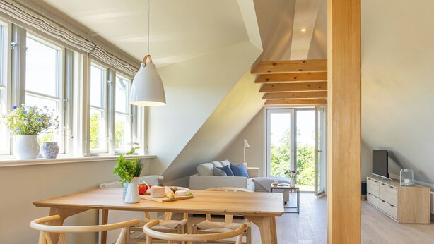 Ett modernt rum i loftstil med en matplats, synliga träbjälkar, stora fönster och en mysig sittgrupp.