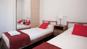 A hotel room with two single beds, each with a red pillow and a white blanket. There is a wooden nightstand with a lamp between the beds.
