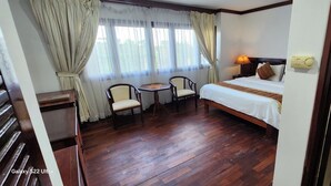 Habitación de hotel con piso de madera, una cama, dos sillas, una mesita y una ventana con cortinas.
