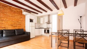 Un salon moderne avec un canapé en cuir noir, un plancher en bois, une table à manger avec des chaises, un coin cuisine avec des meubles blancs et des poutres apparentes en bois au plafond.