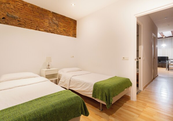 Una habitación con dos camas, una pared de ladrillo, una puerta blanca que da a otra habitación y piso de madera.