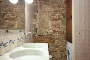 A bathroom with a stone wall, a double sink, and a washing machine. - Elegant Gothic Apartment in El Born   (Barcelona)