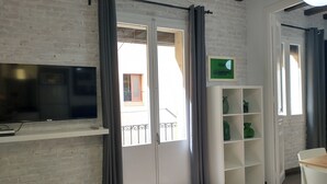 A room with a brick wall, a flat-screen TV mounted on the wall, a white shelving unit with green bottles, and a door with grey curtains. - Lovely flat in Barceloneta near sea (Barcelona)
