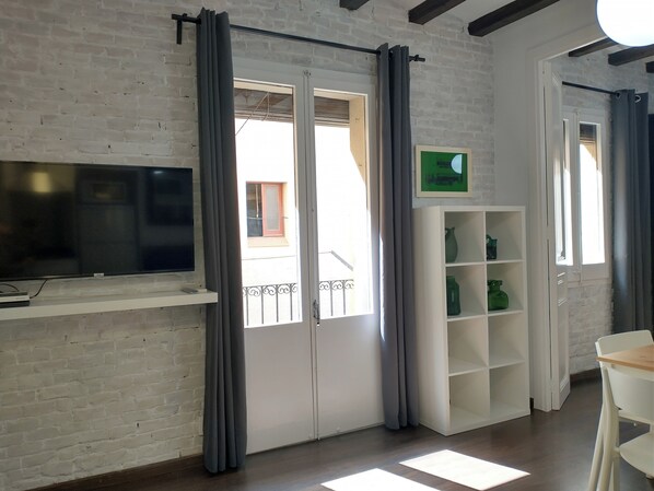 A room with a brick wall, a flat-screen TV mounted on the wall, a white shelving unit with green bottles, and a door with grey curtains.