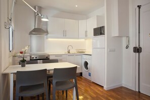A compact kitchen with white cabinets, a microwave, a washing machine, and a dining area with two chairs.