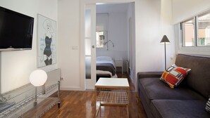Uma sala moderna com televisão de tela plana, um sofá com almofada colorida, uma mesa pequena e uma área de cozinha com micro-ondas e prateleira.