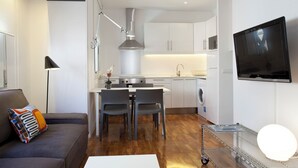 Uma cozinha moderna com armários brancos, coifa de aço inoxidável e máquina de lavar embutida. Há uma área de jantar com mesa e duas cadeiras brancas, e uma TV de tela plana presa na parede.