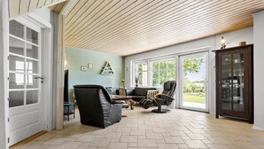 A spacious living area with a wooden ceiling, a black leather sofa, a dining table, and a view of the outdoors through large windows.
