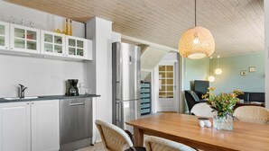 A modern kitchen with white cabinets, a stainless steel refrigerator, and a coffee maker. There is a dining area with a wooden table and wicker chairs.