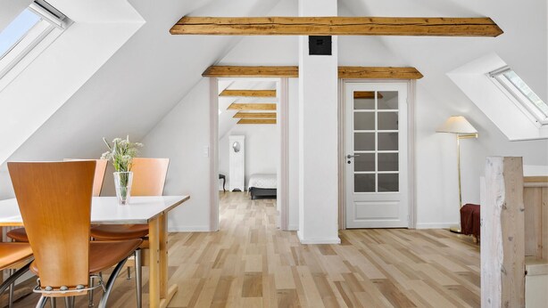 A spacious attic room with wooden beams, a dining area with a table and chairs, and a bed in the background.