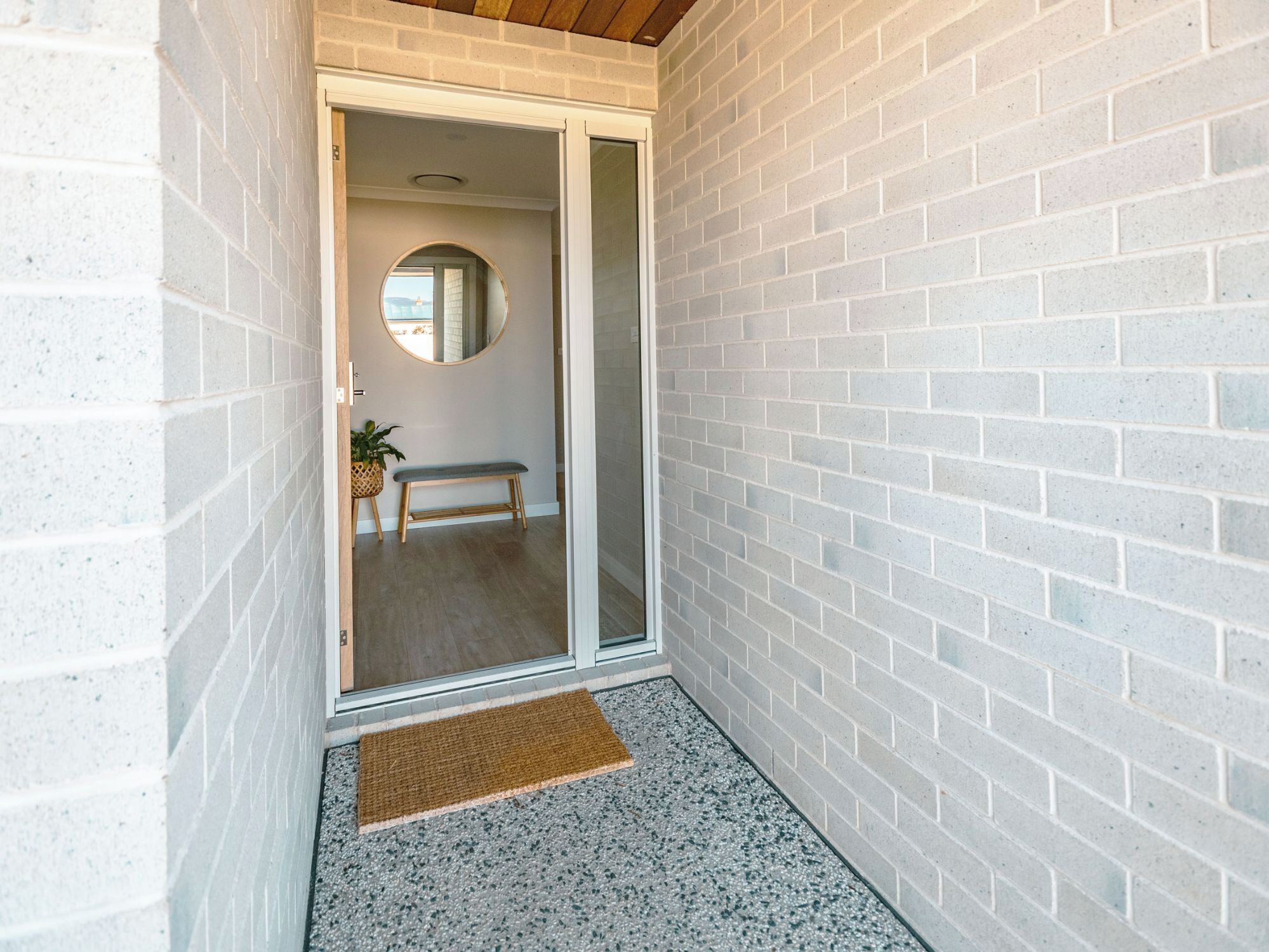 A narrow hallway with a white brick wall, a wooden bench, a small round mirror, and a doormat.