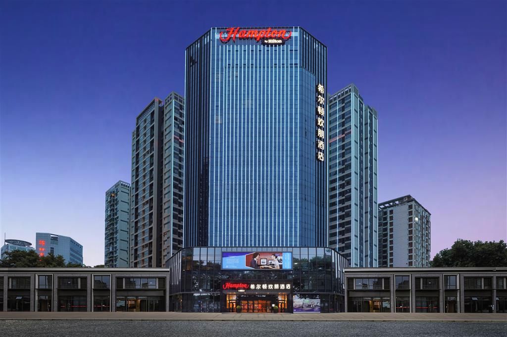 Un hotel moderno con un llamativo letrero de 'Hampton', fachada de vidrio y una entrada central con pantalla digital.