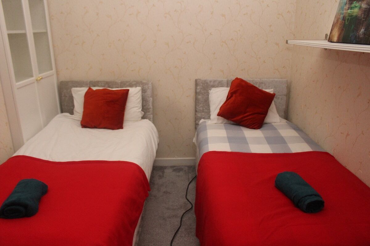 A small room with two single beds, each with a red blanket and pillow. There is a white door on the left and a shelf with a picture on the right.