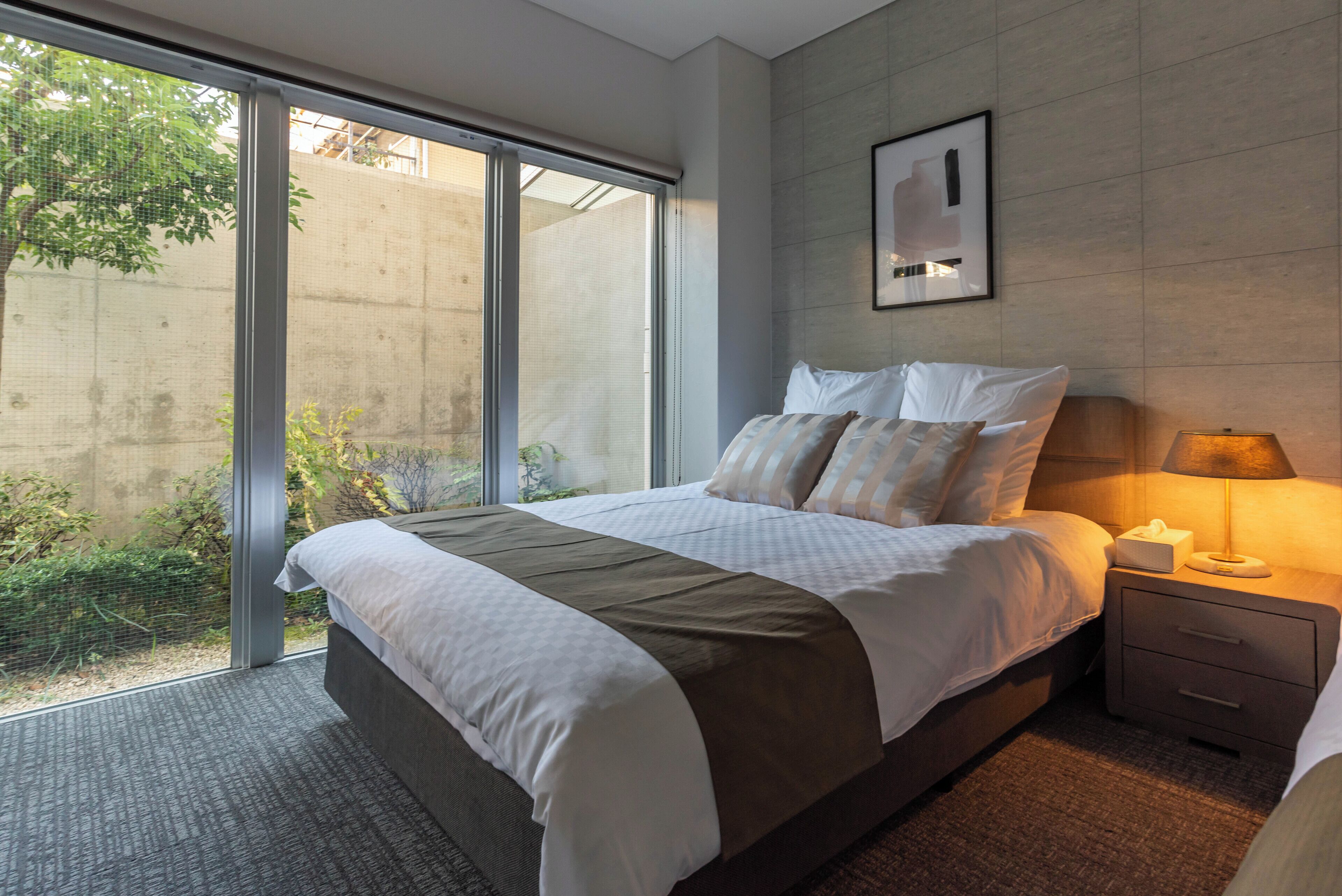A modern hotel room with a large bed, a bedside table with a lamp, and a view of the outdoors through a sliding glass door.