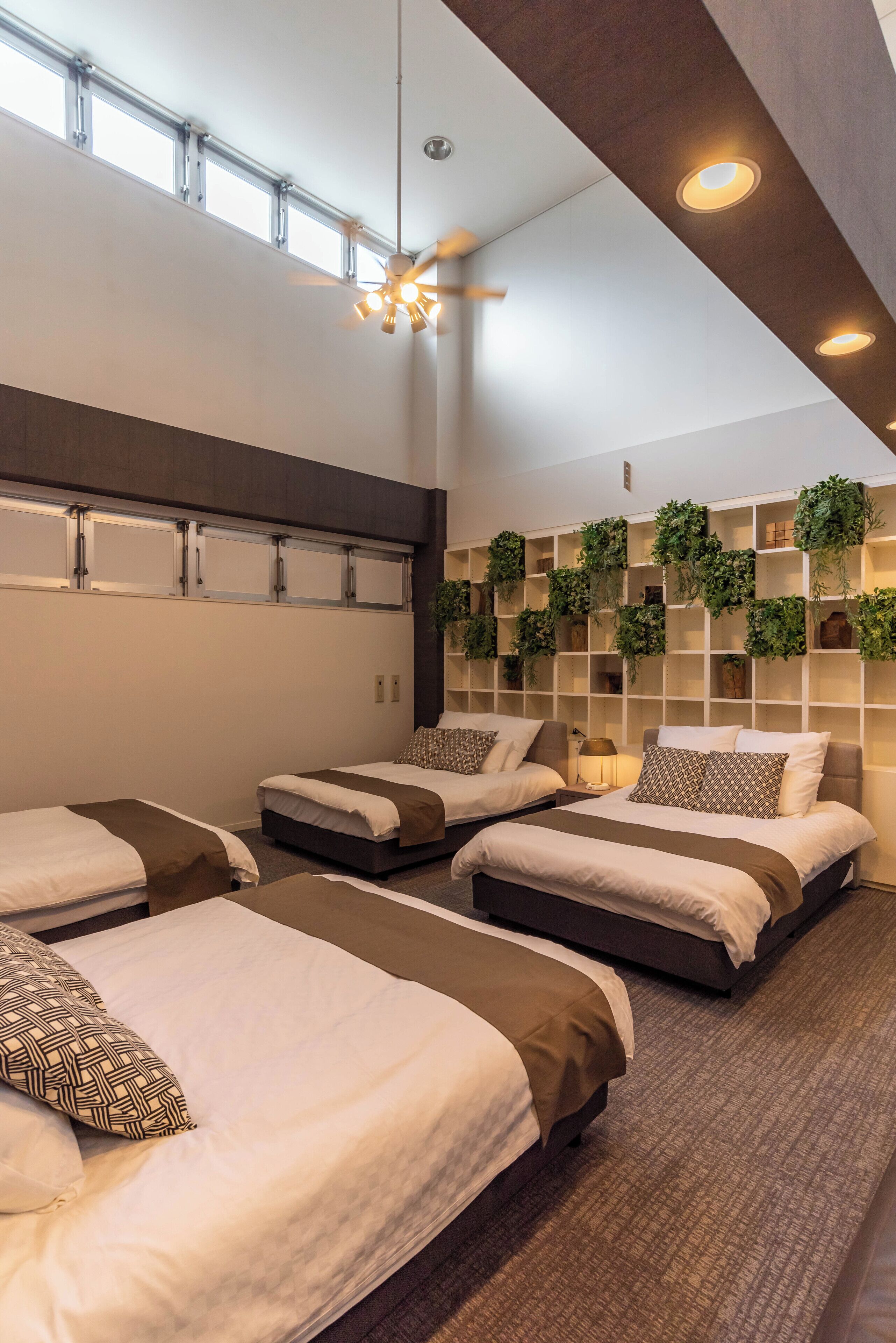 Habitación de hotel con tres camas, ventilador de techo, iluminación empotrada y estante para plantas fijado a la pared.