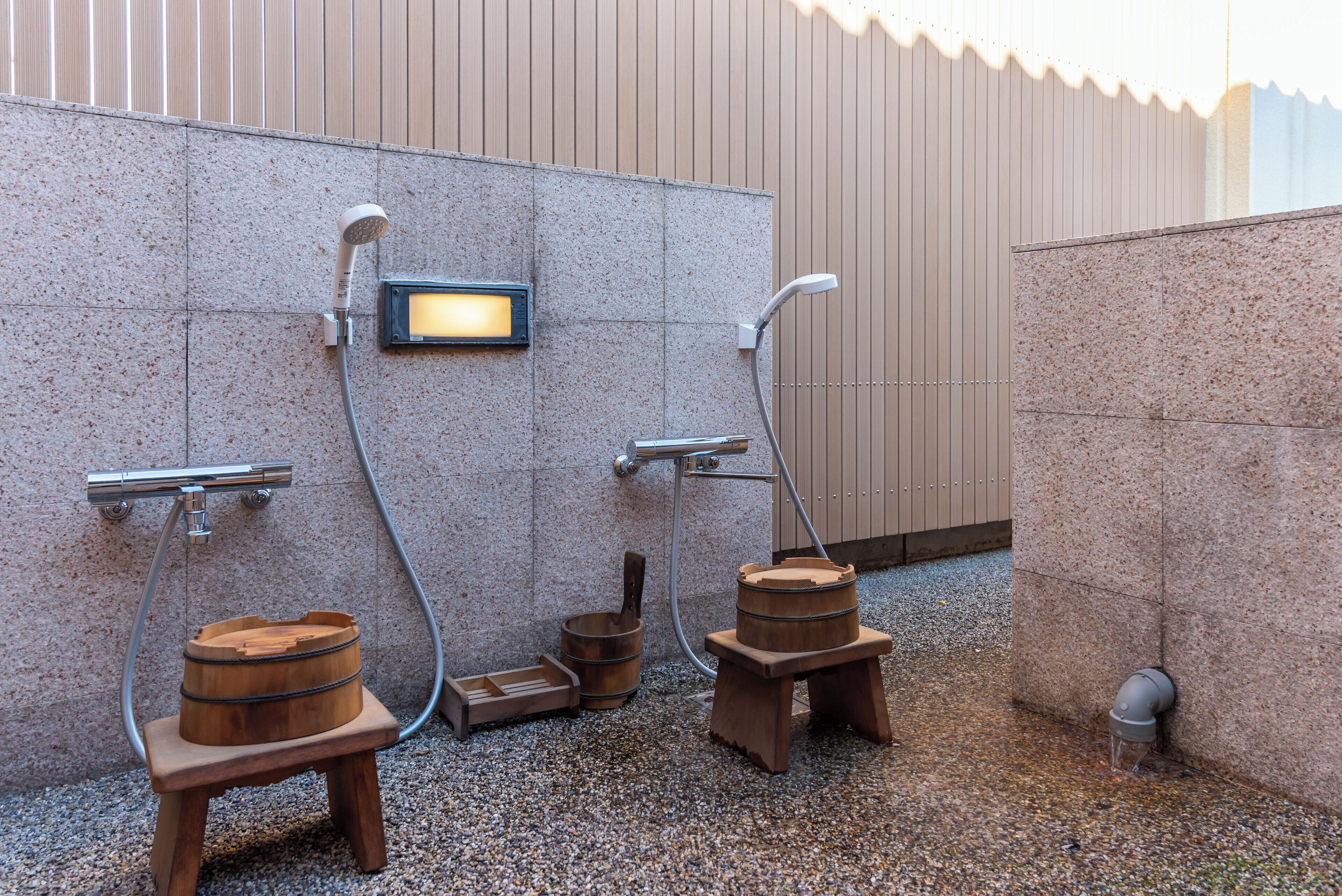 A modern outdoor shower area with two wooden stools, a wooden bucket, and a metal showerhead mounted on a wall.