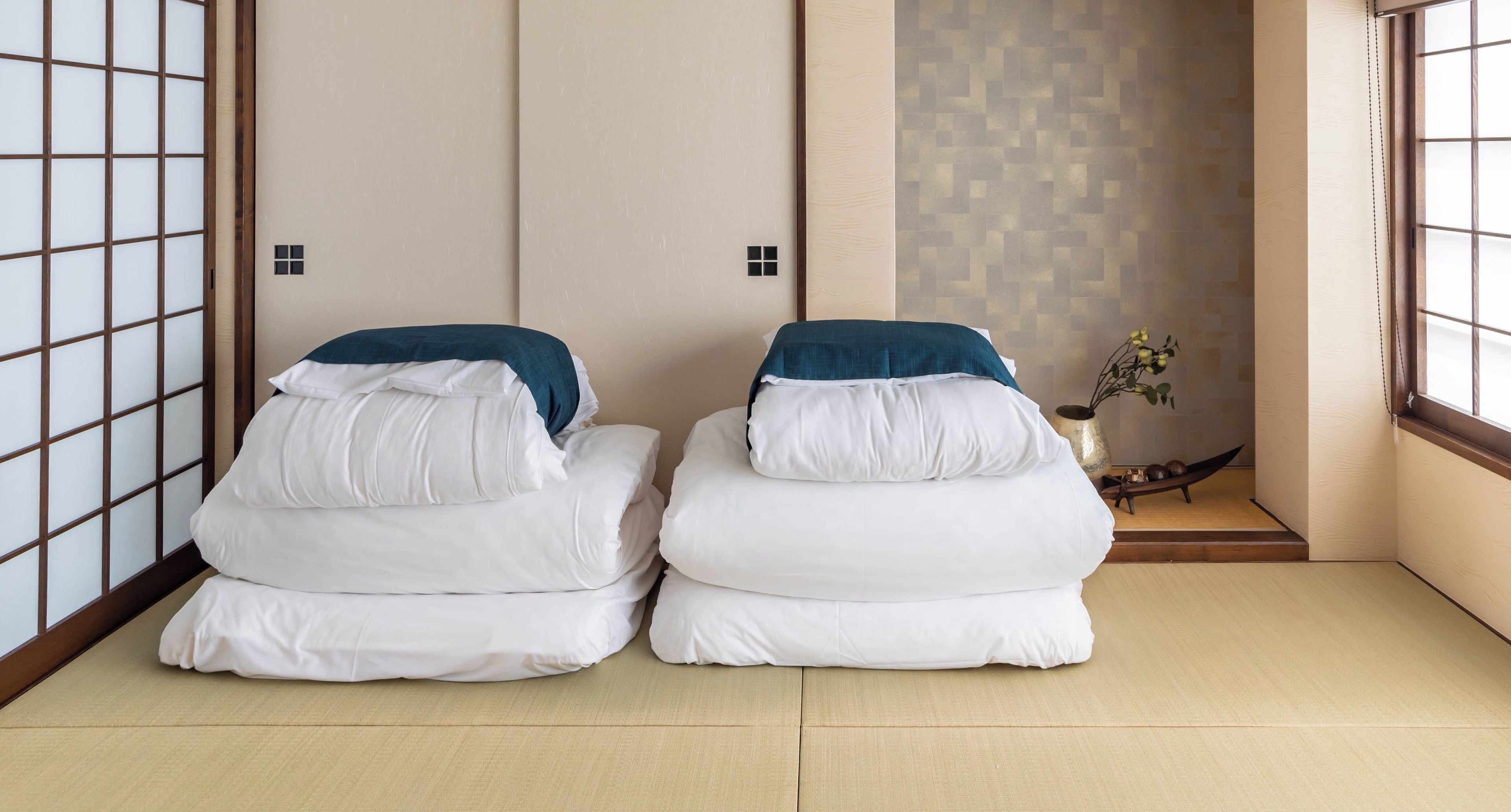 A traditional Japanese-style room with tatami flooring and shoji screens. There are two futons with pillows and a small table with a vase of flowers.