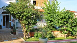 A courtyard with a paved walkway, a brick planter with a tree, and a building with a red roof in the background. - Impeccable 2-bed Apartment in Rab (Rab)