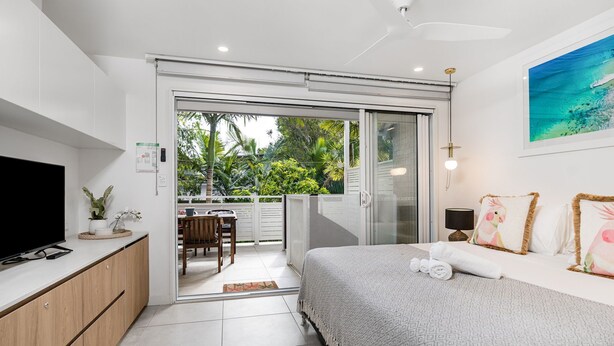 A modern hotel room with a balcony, a TV, a bed with decorative pillows, and a painting of a tropical scene. - Belongil Beach Pad Byron Bay (Byron Bay)
