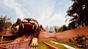 Une grande statue de lion, richement détaillée, située dans un parc, avec un décor rocheux en arrière-plan et un sentier menant jusqu'à elle.
